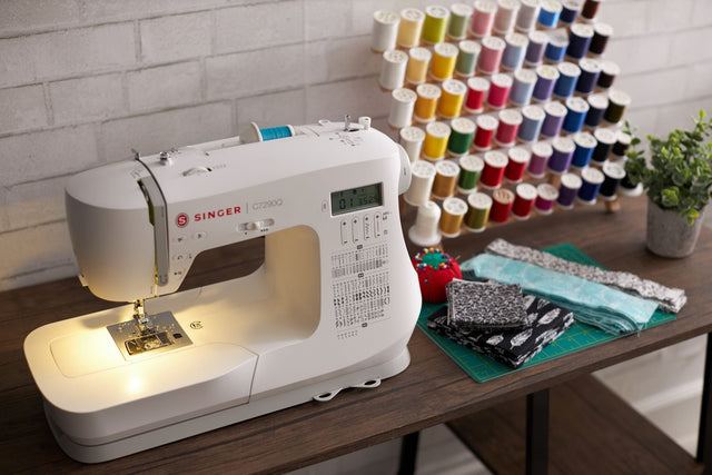 Singer C7290Q sewing machine on a workspace with fabric and thread spools, featured in a comparison with the Brother CS600I.