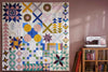 Colorful patchwork quilt with various geometric patterns and vibrant colors, including blue, yellow, pink, and green, displayed on a wall next to a sewing machine and a cabinet filled with fabric and quilting supplies.